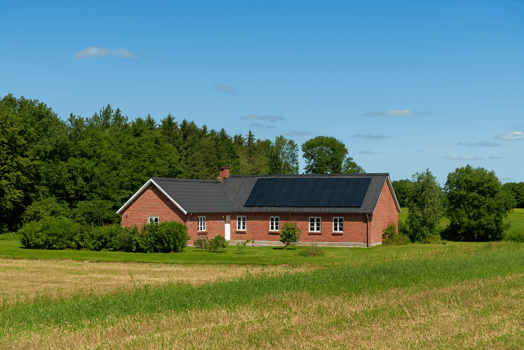 L-længet gård med solceller på taget, omgivet af skov og marker under blå himmel – bæredygtig energiløsning i naturskønne omgivelser